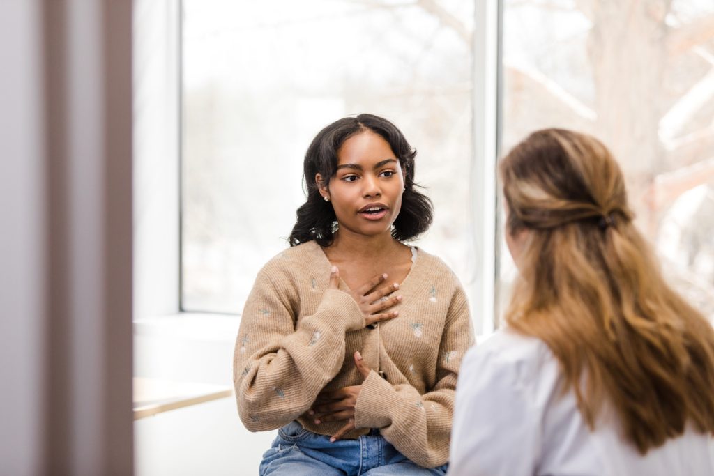 A young woman sits indoors, speaking with a professional who has her back to the camera. The woman gestures with her hand on her chest, appearing engaged in conversation. A large window is in the background.