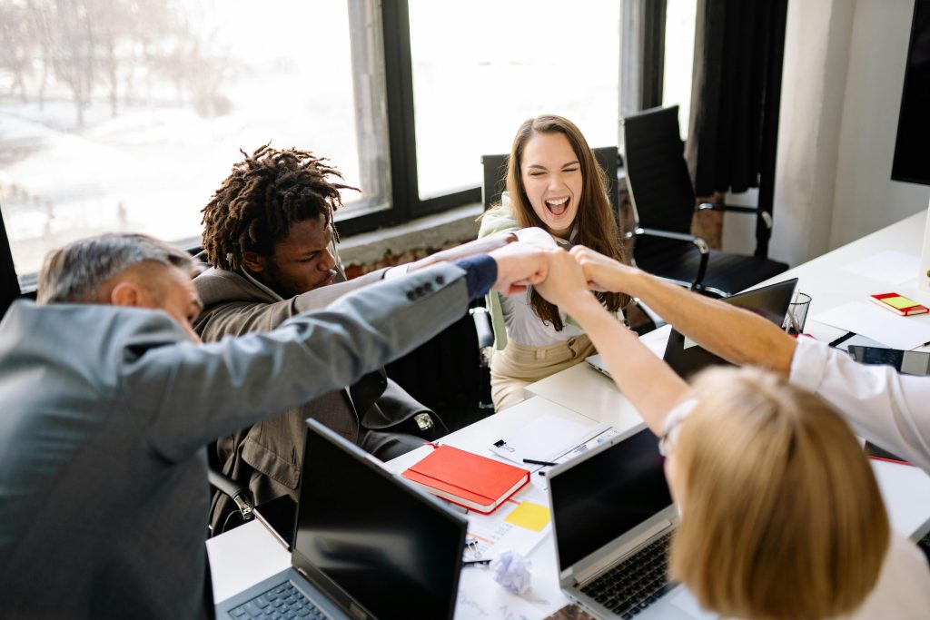 Four colleagues sit around a table in an office, smiling and doing a group fist bump over laptops and notebooks, celebrating teamwork and success.